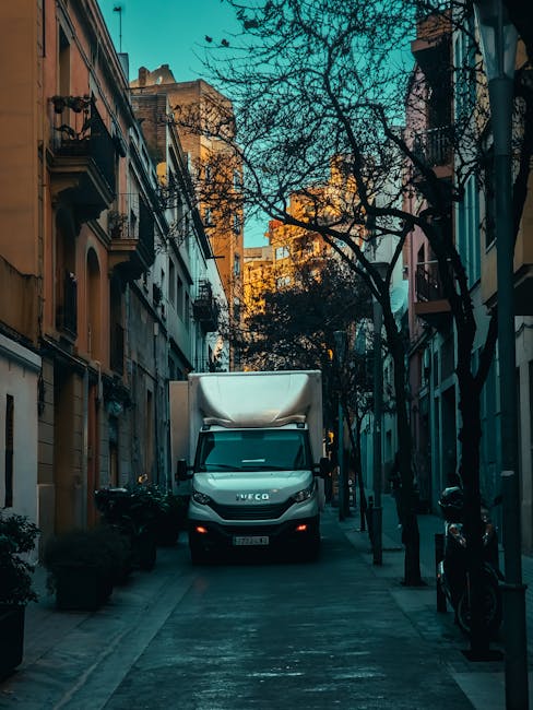 A white Iveco moving van with a covered cargo area is parked in a narrow urban street lined with multi-storey residential buildings featuring balconies and ornate architectural details. The street pavement is made of concrete, and there are black bollards and parked motorcycles along the sidewalk. Bare tree branches stretch overhead, indicating a season of late autumn or early winter. Inside the vehicle's cargo area, visible cardboard boxes, wrapped furniture with plastic and fabric coverings, and packing materials such as bubble wrap are being loaded or unloaded during a home relocation process. The scene is illuminated by natural daylight, with a mix of shadows cast by the surrounding buildings and trees. Ilford Man and Van, a professional removals company, may be coordinating the loading and transport of household items as part of furniture transport and packing and moving services to facilitate an efficient house removal, aligning with the context of avoiding hidden fees in Ilford removals.