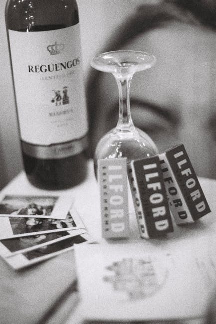 A black-and-white image displays a wine bottle labeled 'Reguengo', partially visible at the left edge on a flat surface, alongside an upside-down wine glass with an empty bowl and slender stem. Near the glass, there are multiple small printed photographs stacked and scattered, with some overlapping. Prominently, three of the photographs feature labels or branded items, including one with the text 'LIFORD' and 'FOR' repeated, possibly part of a promotional or packaging material. The background is slightly out of focus, and the setting appears to be indoors, perhaps in a living room or kitchen, where objects suggest a casual or celebratory atmosphere. This scene could relate to home handling or packing activities associated with house removals or moving preparations, fitting within the scope of domestic relocation processes managed by Ilford Man and Van's removal services.