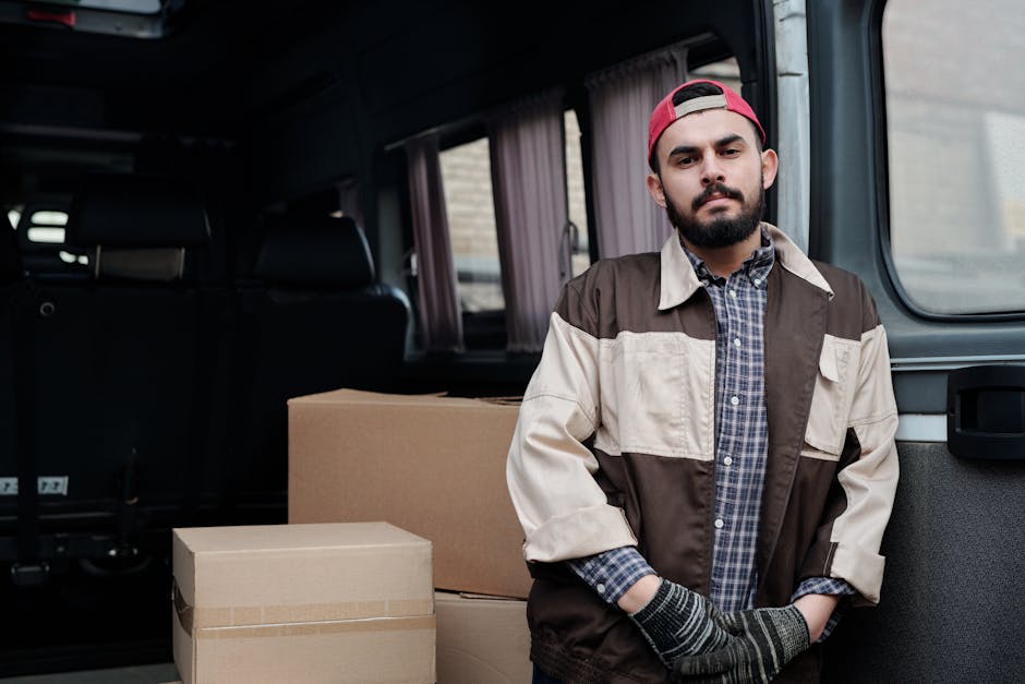 A man with a beard, wearing a red cap, plaid shirt, and a two-tone brown and beige jacket, stands inside a vehicle near the open side door. Behind him, in the interior of the van, there are several packed cardboard boxes, some sealed with packing tape, arranged on the floor. The van's interior has dark seats and light-colored curtains covering the windows. The scene illustrates a home relocation or furniture transport process, with the man appearing to prepare for the loading or unloading phase, which is part of professional removals services offered by Ilford Man and Van. The environment is well-lit, indicating natural daylight streaming through the van windows, emphasizing the logistics involved in packing, loading, and securing household items during a move.