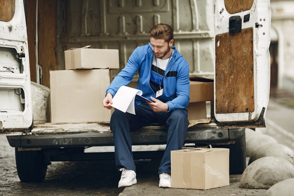 A man in casual clothing, wearing a blue hoodie and dark pants, sits on the edge of an open van tailgate in a warehouse or loading area, surrounded by cardboard boxes of various sizes. He is holding a clipboard or document and appears to be reviewing or checking a list related to a home relocation or furniture transport process. Several boxes are stacked beside him inside the van, which is lined with protective materials such as blankets or padding, indicating a packing and moving operation. The scene is well-lit with natural or artificial light, and the environment features industrial elements like concrete flooring and storage containers. This image illustrates the work involved in professional removals and the planning stage of house moving, aligning with services offered by Ilford Man and Van, who assist with packing, loading, and transport for house removals and relocation services.