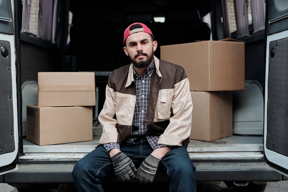 A man with a beard, wearing a red cap, plaid shirt, and a two-tone brown and beige jacket, stands inside a vehicle near the open side door. Behind him, in the interior of the van, there are several packed cardboard boxes, some sealed with packing tape, arranged on the floor. The van's interior has dark seats and light-colored curtains covering the windows. The scene illustrates a home relocation or furniture transport process, with the man appearing to prepare for the loading or unloading phase, which is part of professional removals services offered by Ilford Man and Van. The environment is well-lit, indicating natural daylight streaming through the van windows, emphasizing the logistics involved in packing, loading, and securing household items during a move.