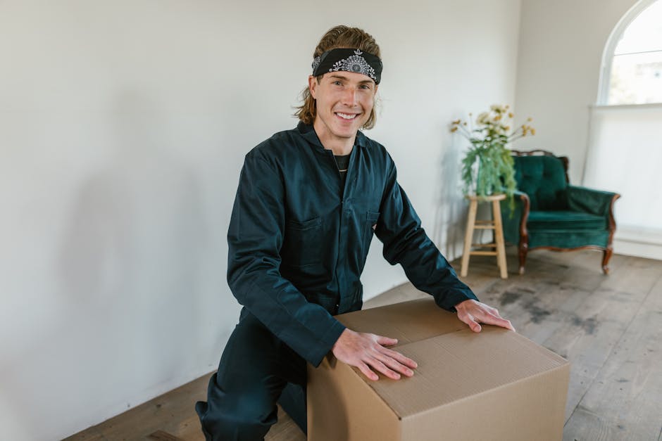 A professional mover from Ilford Man and Van, dressed in a dark uniform, is inside a residential property during a home relocation process. He is smiling and securing a large cardboard box on a wooden floor, preparing it for transport. The room has natural light coming through large, arched windows, with a green velvet armchair and a small wooden side table holding a plant in the background. The environment appears clean and organized, with minimal furniture, highlighting the packing and moving activities involved in house removals. The mover's actions demonstrate careful handling of packing materials, and the scene captures the essentials of furniture transport and loading procedures today, supporting relocation services aligned with house removals on Cranbrook Road IG2, as detailed in the 'Cranbrook Road IG2 house removals best routes and parking tips' guide on ilfordmanandvan.org.uk.