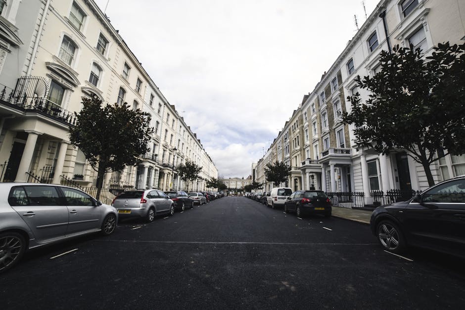 A wide residential street in Ilford, with rows of white, Victorian-style terraced houses on either side. The pavement is lined with parked cars, including a silver hatchback, a grey sedan, and a black SUV, parked parallel to the curb. Several trees with dark green foliage are planted along the sidewalks, providing shade and greenery. The street appears to be in a quiet area with no visible traffic or pedestrians. The sky is partly cloudy with patches of blue. The scene depicts a typical setting for house removals or furniture transport, where vehicles are positioned close to the property entrances for loading or unloading, making it relevant for discussions on house removals, parking, and moving logistics. Occasionally, Ilford Man and Van's services could assist with the house moving process in such environments, ensuring efficient loading and safe transport of household items.