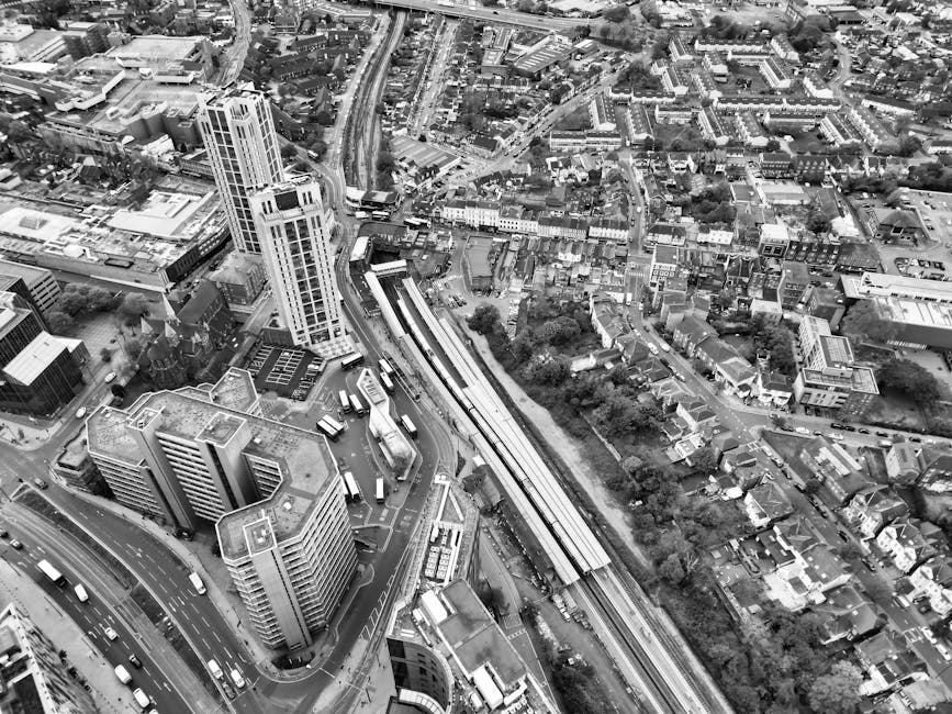 An aerial black-and-white photograph capturing an urban area with tall office buildings and residential houses. The image shows a busy cityscape with multiple roads, including a curved multi-lane roadway at the bottom left and a railway line running diagonally across the scene, near a train station platform. Several buses and cars are visible on the roads, and the train station has a large, elongated platform with trains parked alongside. Surrounding the transportation infrastructure are various buildings, some with flat roofs, and densely packed houses with narrow streets and green spaces in the background. Natural light illuminates the scene, highlighting the detailed layout of the city. This image is related to house removals and relocation logistics, showcasing the complexity of urban transport and the potential environment for packing, moving, and furniture transport conducted by services like Ilford Man and Van.