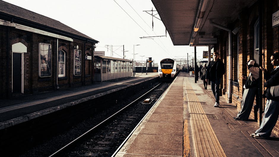 A view of Ilford train station platform during daytime with several passengers waiting for a train. The platform is made of concrete with tactile paving near the edge to assist visually impaired travelers. On the right side, there are people standing, some carrying bags or luggage, dressed in casual clothing suitable for travel. The station building, constructed from brick with advertisements displayed in window frames, is visible on the left side. Overhead, there are metal canopies providing shelter, and railway tracks run along the platform. In the background, a modern, white-and-yellow train is approaching or has just arrived, with overhead electrical lines visible above the tracks. The scene is illuminated by natural daylight, casting shadows on the platform, and the overall setting reflects a busy railway station environment, relevant to train travel and station logistics, which are part of house relocation or moving logistics services offered by Ilford Man and Van.