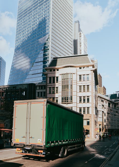 A large green cargo truck with an enclosed box body is parked on a city street in front of modern high-rise office buildings and older stone architecture. The truck is positioned on the road adjacent