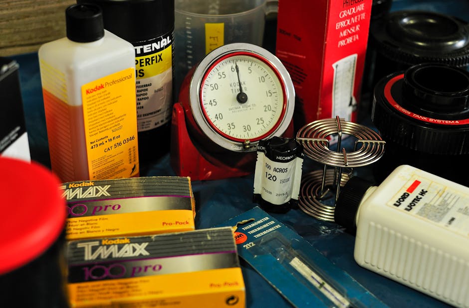 A close-up view of various photographic and electrical items arranged on a surface, including two rolls of Kodak film labeled 'TWAX 100 pro' and 'MAX', a white bottle of Kodak Professional fixer, a vintage red kitchen scale with a circular dial showing grams and ounces, a large white plastic container, several black and red camera lenses or filters, a metal coil spring, a small black and white capacitor, and a blue card with some text. The items are positioned in front of a blurred background, possibly inside a home or moving environment, with some items overlapping each other. The scene features different textures such as plastic, metal, and cardboard, and suggests packing or preparation for transport related to a home relocation or moving process, which Ilford Man and Van might handle as part of their removals services.