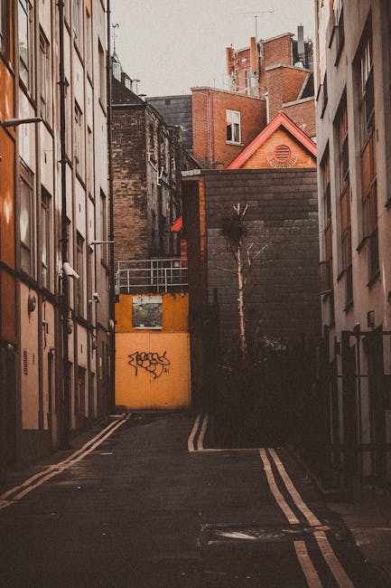 A narrow street in an urban residential area with closely positioned multi-storey buildings on each side, constructed from brick and concrete, some of which feature balconies, windows, and fire escapes. The street has double yellow lines along the edges and appears to be an access route for vehicles and moving activities. At the end of the street, a yellow metal gate with graffiti and a small door is visible, leading to a back alley or courtyard. The surrounding buildings include various flat roofs and pitched roofs with red tiles, and some have ventilation ducts or chimneys. The lighting suggests late afternoon or early evening, casting warm tones across the scene. This setting exemplifies an access problem for house removals, often addressed by companies like Ilford Man and Van during home relocation and furniture transport operations in tight urban spaces.