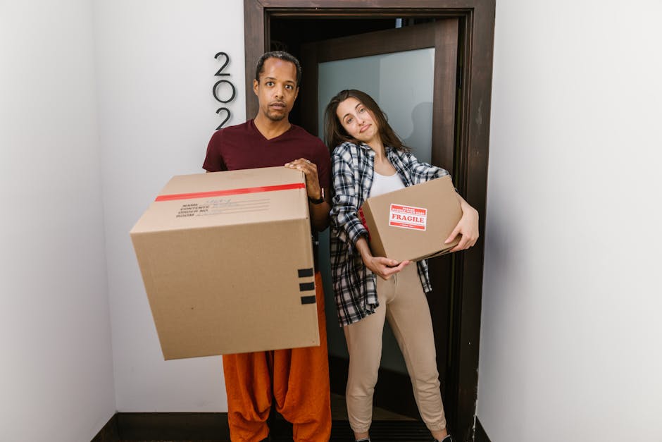 A man and a woman standing inside a doorway with a dark wooden frame, each holding a cardboard box in preparation for home relocation. The man is wearing a maroon t-shirt and orange trousers, holding a large medium-sized box with a red and white shipping label. The woman is dressed in a plaid shirt over a white top and beige trousers, carrying a smaller box marked with a red 'Fragile' sticker. They are positioned on a light-colored hallway floor, with an interior room visible behind them. The background includes a white wall with black house number plaques, indicating a private residence. Several packing materials such as plastic wrap or blankets are not visible, but the focus is on their process of packing and moving household items. This image exemplifies the loading process during furniture transport and home moving services, with a natural indoor environment and clear focus on packing and handling boxes, aligning with the topic of same-day removals and urgent moves by Ilford Man and Van.
