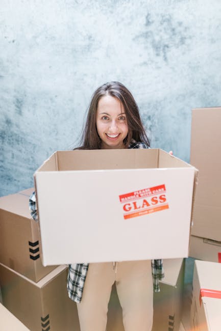 A young woman with long brown hair and a cheerful expression is standing indoors against a light blue textured wall. She is holding an open cardboard moving box labeled 'FRAGILE GLASS' in red text, with her hands supporting the sides of the box. Behind her, there are several other cardboard boxes of various sizes, some sealed with packing tape, stacked on the floor. One box appears to be ready for transportation, suggesting preparations for a home relocation or furniture transport. The scene is well-lit with natural or soft artificial lighting, emphasizing the clarity of the objects and the environment. The setting indicates a packing and moving process, likely part of a professional removal or moving service, as offered by Ilford Man and Van, specializing in same-day removals and urgent moving solutions within Ilford.
