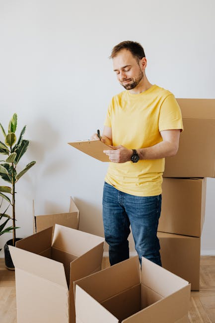 A man wearing a yellow t-shirt and jeans stands indoors in front of a white wall, surrounded by open cardboard boxes and packing materials. He is holding a clipboard and pen, appearing to review or check off items as part of the home relocation process. To his right, there are several packed boxes stacked against the wall, ready for moving. A potted plant with green leaves is visible on the left side of the image, adding a touch of natural décor to the space. The scene depicts a professional packing and moving preparation, with controlled indoor lighting highlighting the details of the boxes and the man's focused expression. This setting reflects the packing and loading phases typical of house removals, which [COMPANY_NAME], such as Ilford Man and Van, provides as part of their furniture transport and moving services, supporting smooth and organized home relocation within the Ilford area.