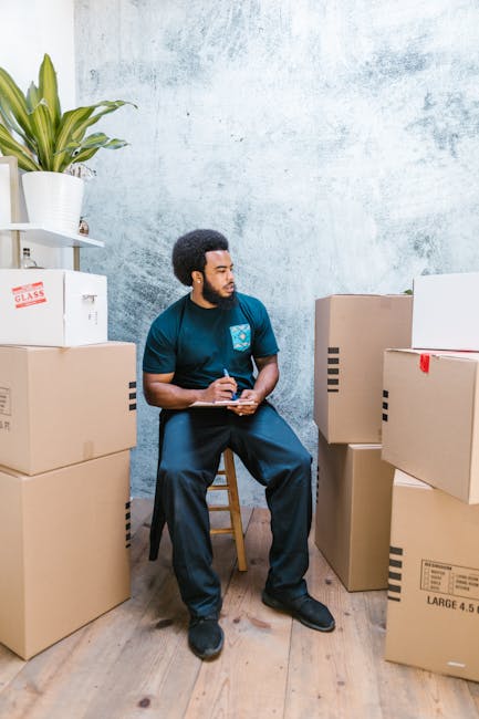 A person wearing a yellow T-shirt and blue jeans stands inside a room surrounded by several large cardboard boxes, some of which are open and empty while others are stacked against the wall. They are holding a clipboard in one hand and using a black marker with the other to record information, likely related to a home relocation or packing process. The room has wooden flooring and is decorated with a potted plant with broad green leaves placed on the floor near the boxes. The lighting is natural, and the scene depicts a stage of packing and moving preparation typical of professional removals services like those provided by Ilford Man and Van, involving furniture transport and loading equipment.
