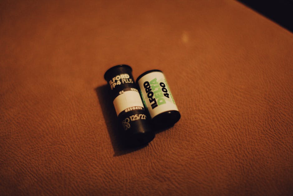 Close-up of two camera batteries resting on a textured brown surface, likely part of packing materials for a house removal. One battery is labeled for a Ford FP Plus camera with a black and white design, while the other is a green and white branded battery, possibly for another electronic device. The batteries are positioned side by side, with subtle shadows indicating ambient lighting. This image illustrates packing and inventory organization during a home relocation, as carried out by Ilford Man and Van, who specialise in furniture transport and packing services for flats and properties near Valentines Park, Parkside. The scene emphasizes careful handling and preparation of electronic equipment alongside moving furniture, boxes, and other household items in the process of a comprehensive removal service.